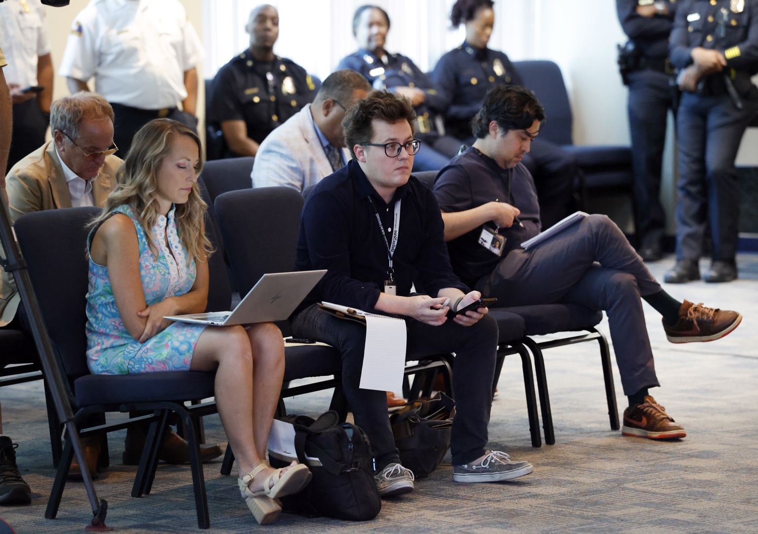 Chase Rogers, seated at center with glasses and a notebook, reporting at a briefing.